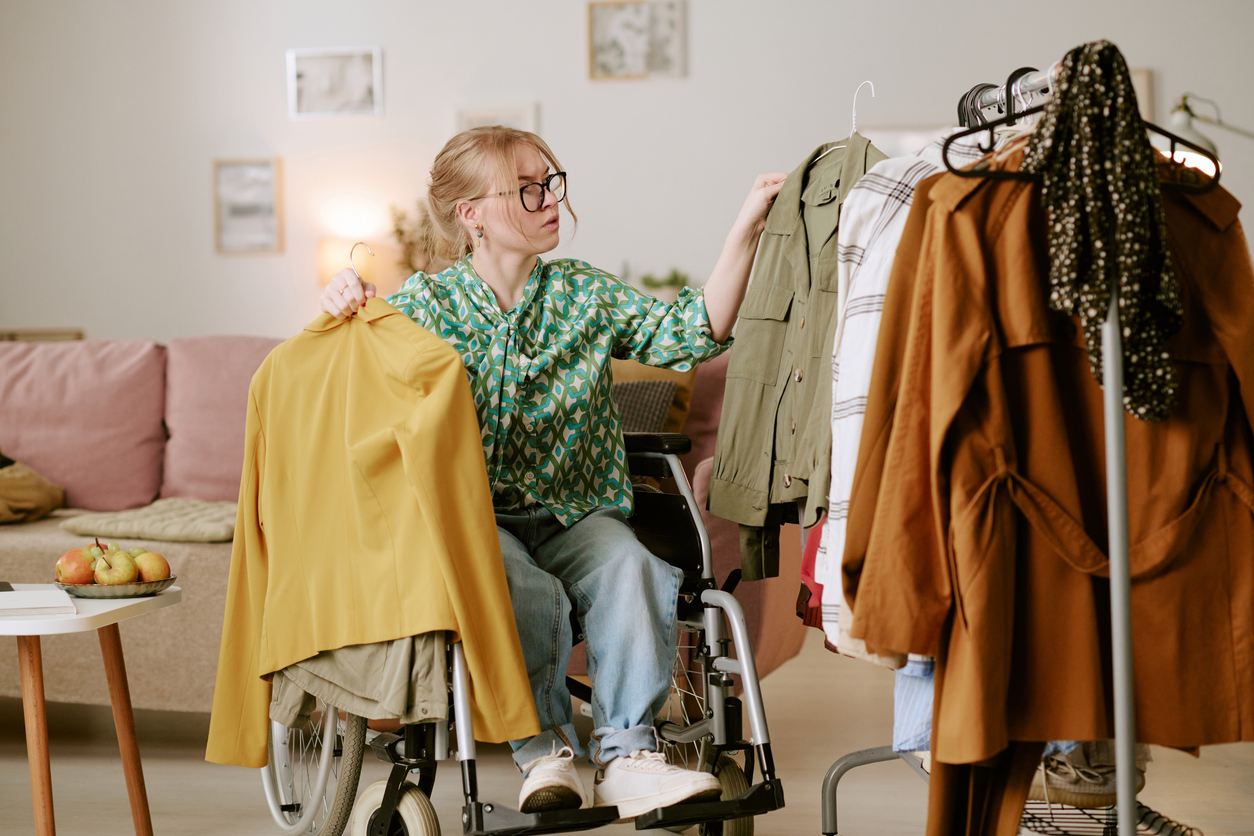 Caucasian young adult woman with disability sitting in wheelchair selecting clothes from rack, holding jacket in one hand, choosing outfit in modern living room setting