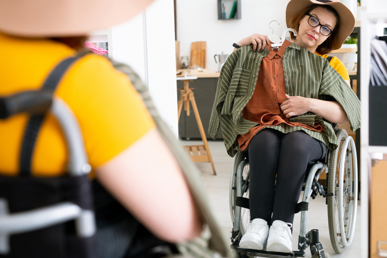 Young disable woman sitting in wheelchair in front of mirror while trying on new hat and smart casualwear that she bought in online boutique